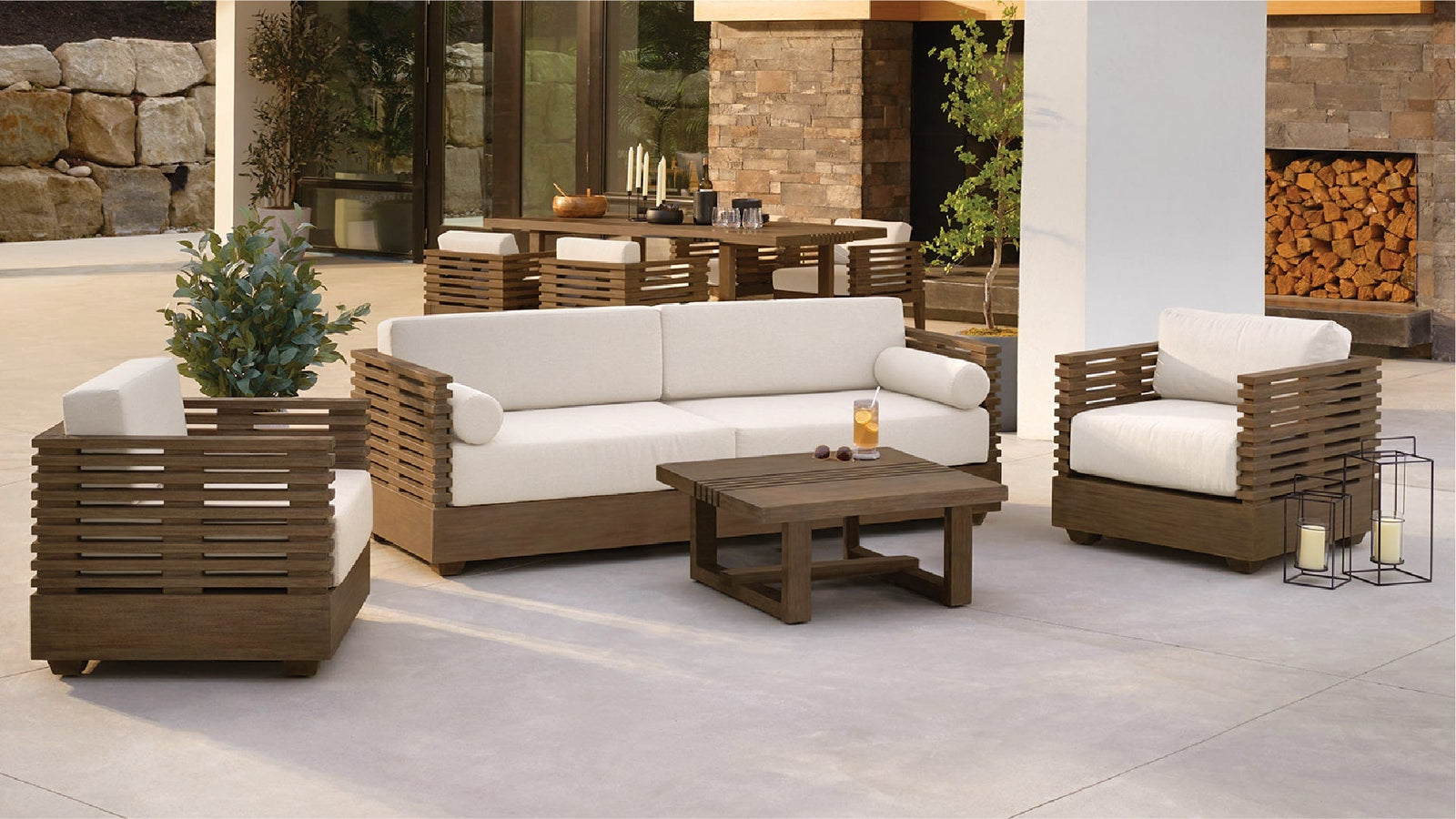 A patio set made of dark brown wood/timber, with off-white cushions, is arranged on a light beige/gray patio floor. 
